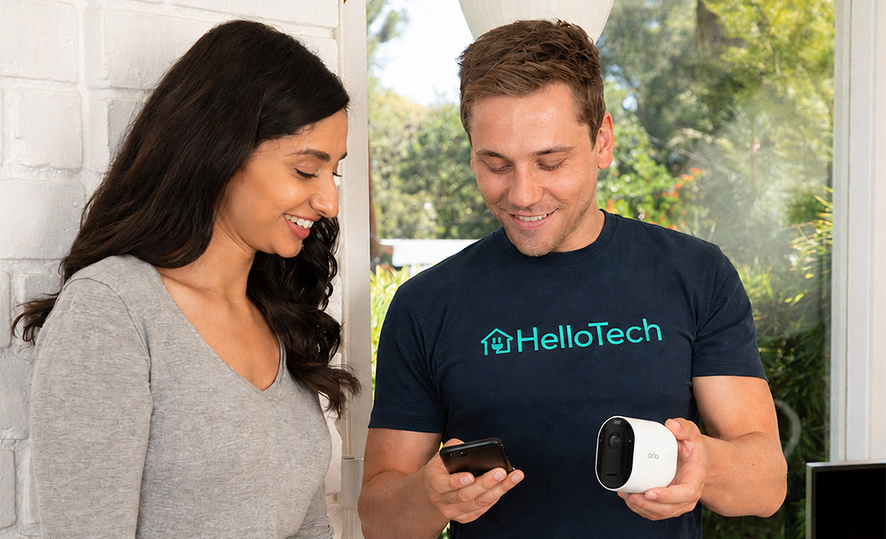 A man wearing a navy HelloTech t-shirt holds a Arlo Pro Security Camera (6th Gen) while looking at a phone, standing next to a woman in a gray long-sleeve top in a bright indoor setting.