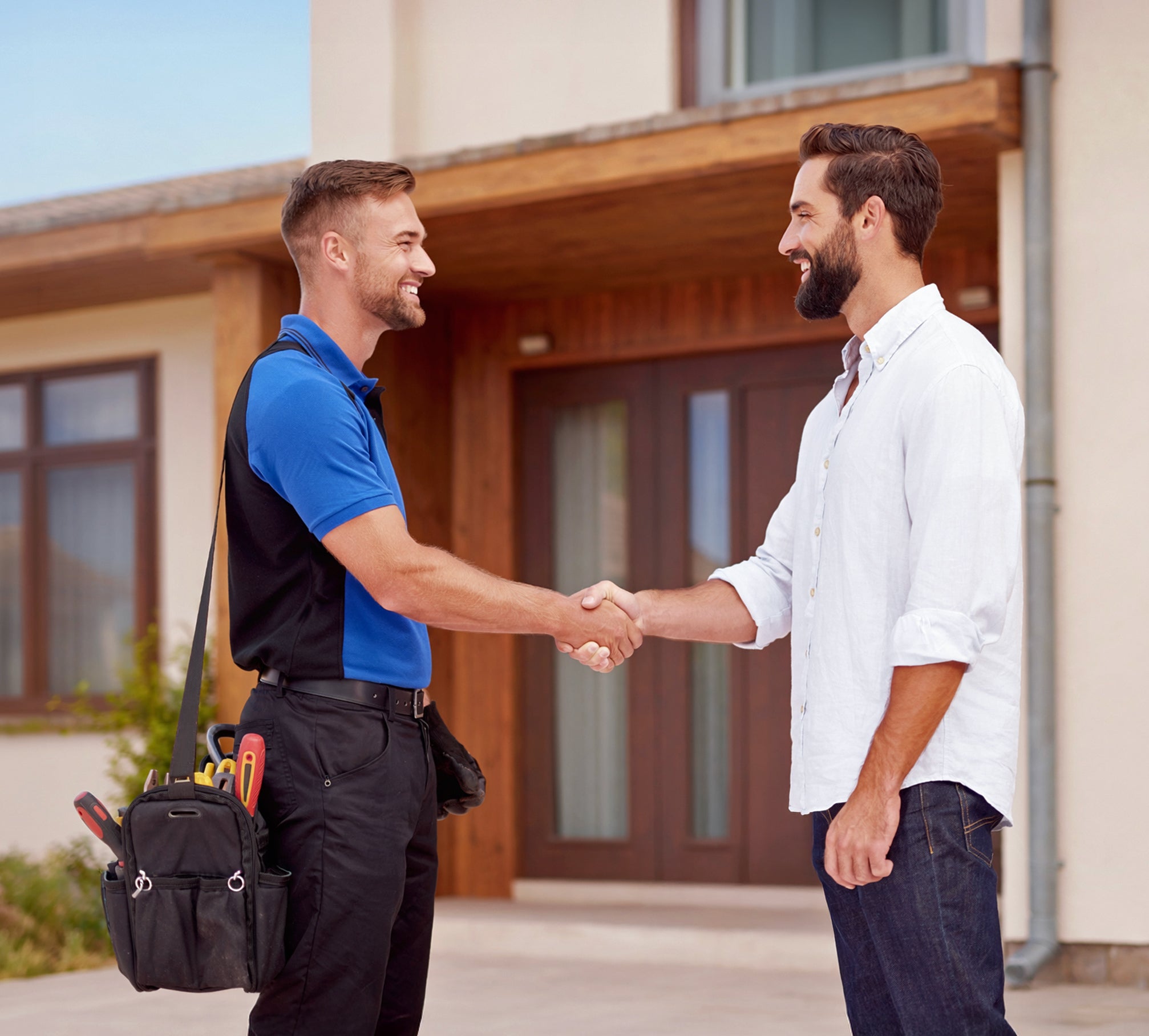 Two men shaking hands in front of a house, one carrying tools.