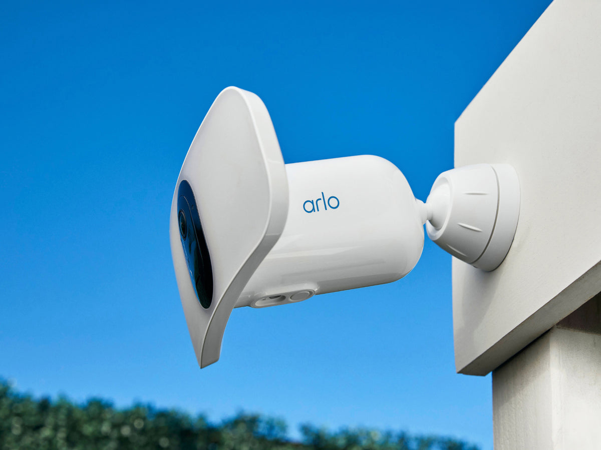 A white Arlo wireless floodlight camera shown mounted on a white post against a bright blue sky background.