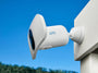 A white Arlo wireless floodlight camera shown mounted on a white post against a bright blue sky background.