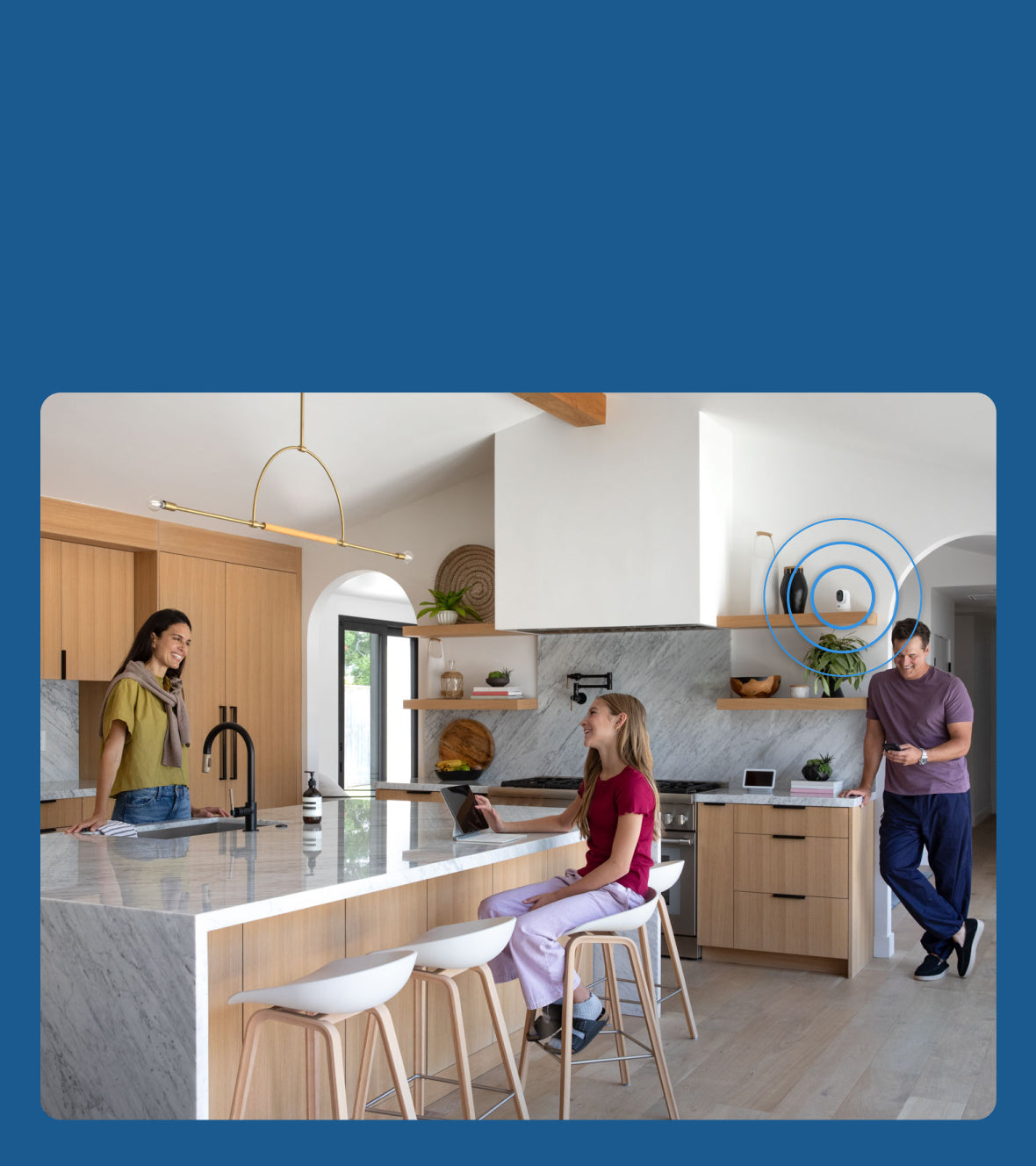 Modern kitchen with people interacting, featuring a Arlo Essential Pan Tilt Indoor Security Camera on the shelf.