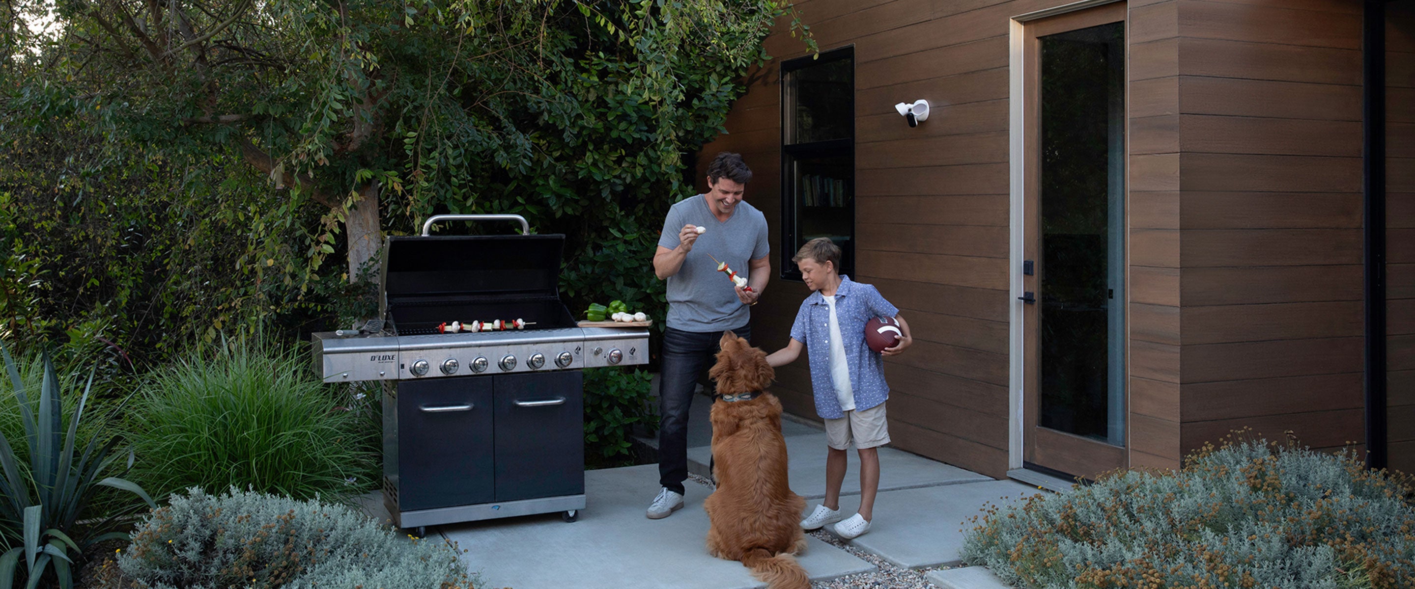 A father, son, and dog outside BBQing with the Arlo Wired Floodlight mounted on the outside of a house