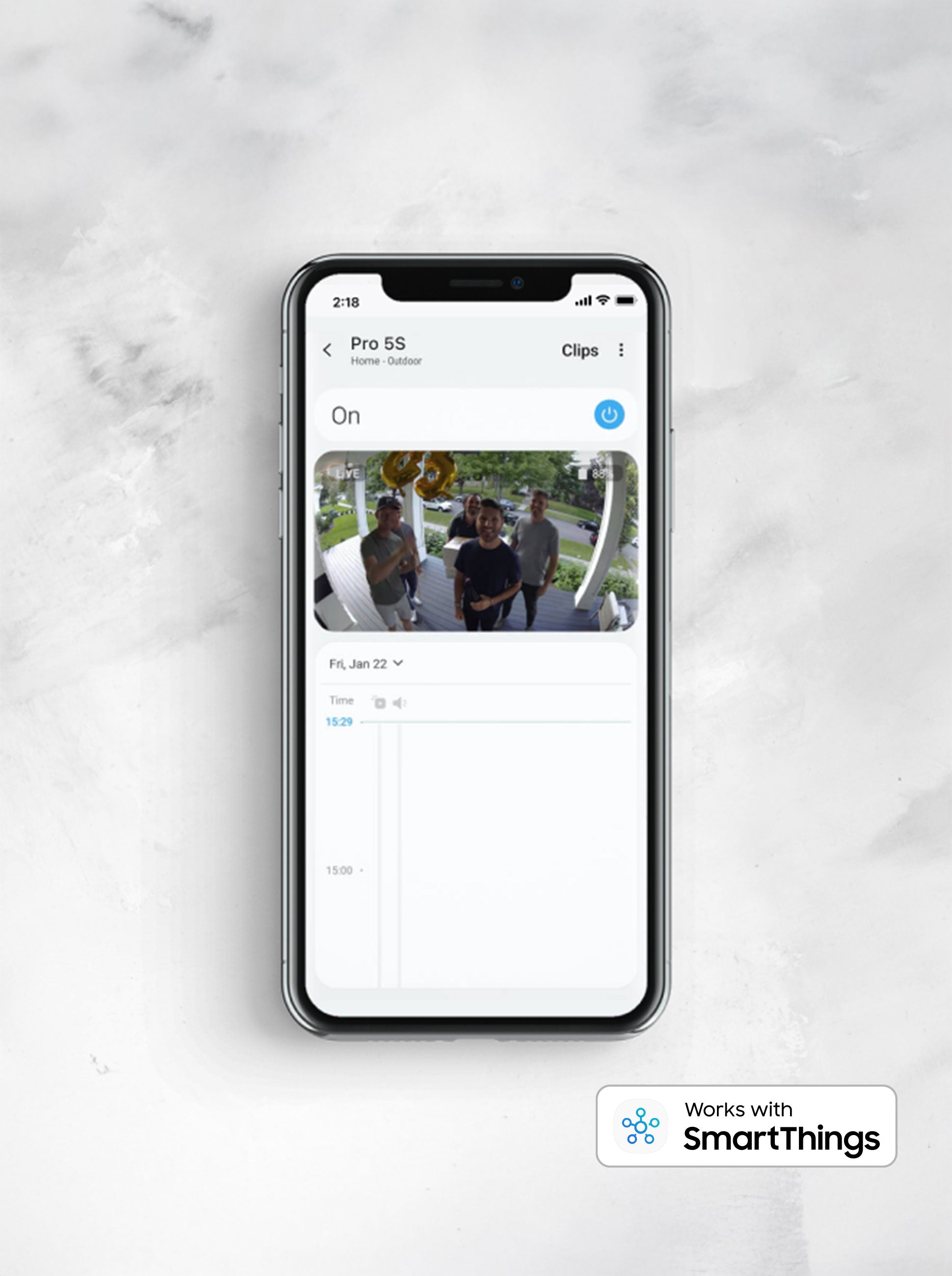 A mobile device placed flat on a marble counter, displays a feed from an Arlo security camera, showing a group of individuals at the front door, smiling, with balloons and gifts. The camera is compatible and pairs with Samsung SmartThings mobile devices for convenient visual & voice control.